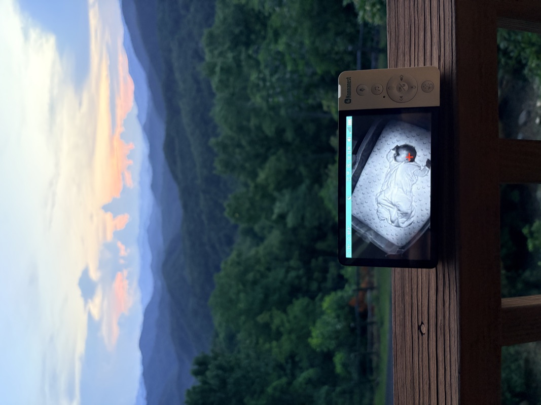 The Nonnanest handheld resting on a deck railing at dusk, showing a sleeping baby, with mountain ridges in the distance.